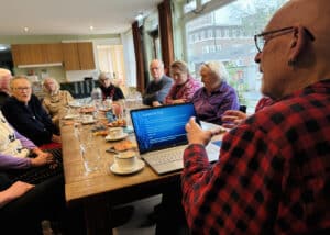 Een man zit aan het hoofd van een lange tafel en legt iets uit aan de deelnemers aan weerszijden van de tafel die aandachtig luisteren